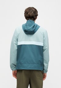 Άνδρας που φοράει μπλε μπουφάν με κουκούλα σε μπλοκ χρωμάτων και χακί παντελόνι, γυρισμένος με την πλάτη προς το φωτεινό, απλό φόντο.