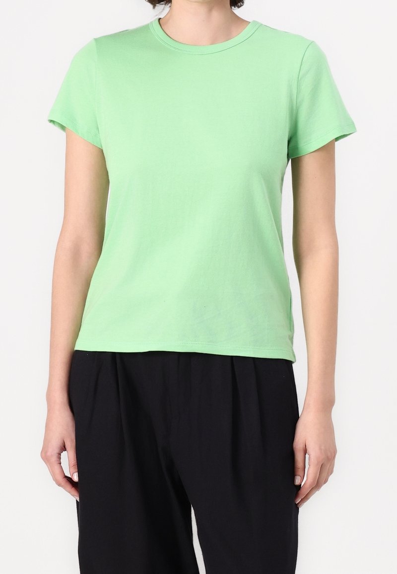 Personne portant un t-shirt vert clair uni à manches courtes et un pantalon noir ample, debout devant un fond blanc.