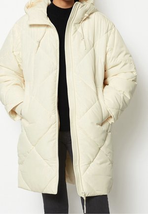 Persoon draagt een lange, gebreide winterjas in off-white met capuchon, zijzakken en rits, over een zwarte trui en donkere broek.