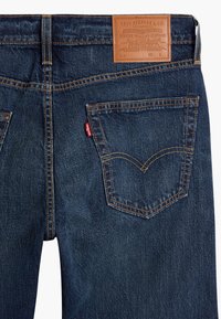 Jeans de mezclilla azul oscuro con un parche de cuero en la cintura. Presenta clásicos bolsillos traseros con costuras naranjas en contraste y una etiqueta roja.