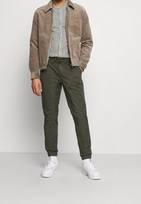 Braune Wildlederjacke, graues gestreiftes Hemd, olivgrüne Hose und weiße Sneakers. Die Jacke hat einen Kragen und eine Fronttasche; die Hose besitzt elastische Bündchen.