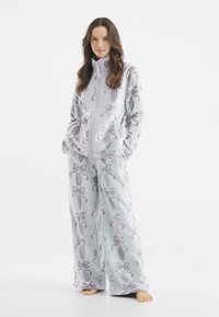 Lyseblåt fleece loungewear sæt med jakke med lynlås og lommer samt brede bukser, begge prydet med et båndmønster.