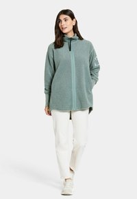 Fleece zip-up tröja i dämpad grön, med hög krage, långa ärmar och sidofickor. Matcherad med vita byxor och ljusa sneakers.