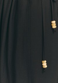 Černá tkanina se stahovací šňůrkou, na jejímž konci jsou ve skupinách po třech navlečené béžové dřevěné korálky.