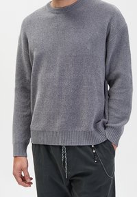 Grauer Strickpullover mit strukturiertem Muster, Rundhalsausschnitt und geripptem Saum sowie Bündchen, getragen über dunklen Hosen mit Akzentriemen.
