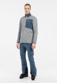 Grijze fleecejack met een marineblauwe borstzak en gouden rits. Gecombineerd met blauwe broek met zwarte accenten, ideaal voor buitenactiviteiten.