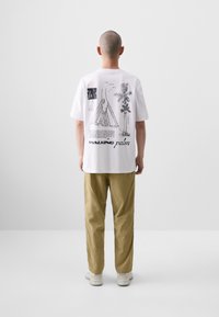 White short-sleeve t-shirt featuring graphic prints on the back, including sketches and text. Paired with khaki pants and white shoes.