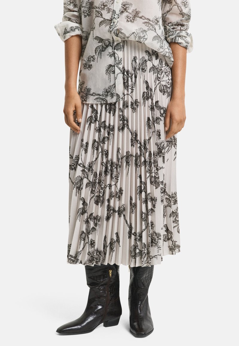 Jupe plissée gris clair avec un motif floral noir ; associée à des bottines noires brillantes avec une finition texturée et des détails de zip doré.