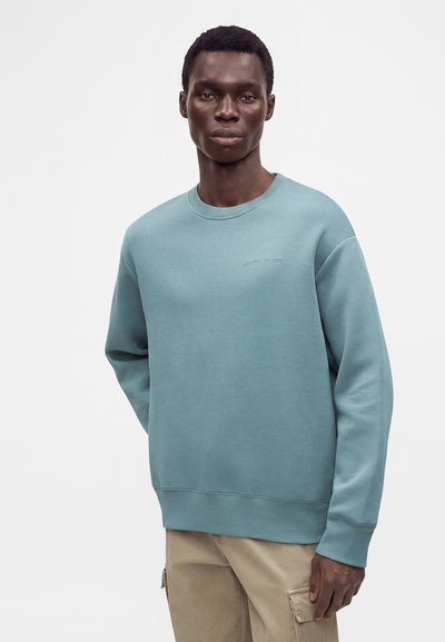Homme portant un sweat-shirt bleu clair à col rond et un pantalon cargo beige sur un fond blanc uni.