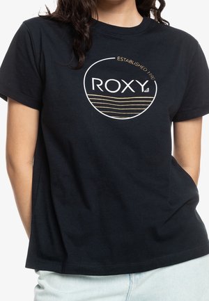 Person iført en sort T-shirt med en rund "ROXY"-logo og teksten "ESTABLISHED 1990" over bølgede gyldne linjer på brystet.