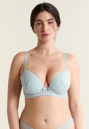 Jonge vrouw met donker haar, draagt lichtblauw kanten beha en bijpassende slip, tegen een effen beige achtergrond, kijkt naar voren.