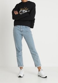 Person står med armarna i kors iförd en svart Nike-hoodie, ljusblå uppvikt jeans och vita sneakers framför en enfärgad bakgrund.