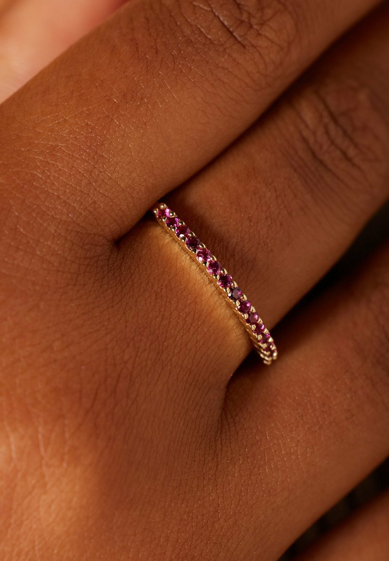 Gouden ring met een rij afwisselend paarse en roze edelstenen, gezet in een delicate band, rustend op een hand met een donkere huid.