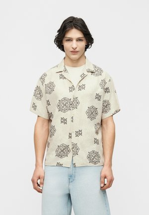 Młody mężczyzna o ciemnych, falowanych włosach, ubrany w beżową koszulę na krótki rękaw z ciemnymi kwiatowymi wzorami oraz jasnoniebieskie dżinsy.