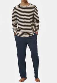 Muž oblečený v béžovom a tmavomodrom pruhovanom tričku s dlhým rukávom a tmavomodrých nohaviciach, stojaci bosý s rukami v vreckách.