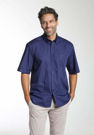 Homme aux cheveux courts et foncés et barbe, portant une chemise bleue marine à manches courtes avec boutons et un pantalon beige clair, mains dans les poches, souriant.