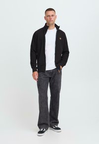 Ung mann står iført en svart glidelåsjakke over en hvit t-skjorte, falmede svarte jeans og svarte joggesko med hvite såler.