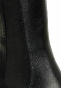 Botín de cuero negro de tobillo con una textura suave y paneles laterales elásticos para facilitar su uso. El diseño incluye un brillo sutil y una forma estilizada.