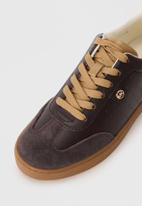 Dark brown sneaker with tan laces, suede toe, perforated side, and gold logo accent on light brown rubber sole.