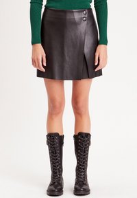 Mini-jupe en cuir noir avec une fente à l'avant, deux boutons sur les côtés, et une texture lisse ; assortie à des bottes noires lacées jusqu'aux genoux.