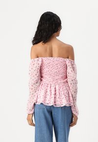 Pink Off-Shoulder-Bluse mit geschopptem Oberteil und langen, puffigen, transparenten Ärmeln, verziert mit kleinen Blumenmustern. Getragen mit blauen Jeans.