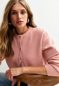 Mujer con cabello largo y rubio, usando pendientes de aro plateados y un cardigán abotonado de un suave color rosa, sentada en una silla de madera.