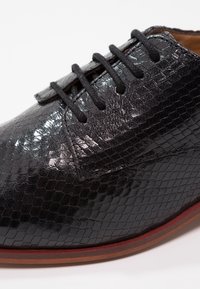 Chaussure noire à lacets en cuir texturé avec un motif hexagonal. Présente une finition lisse et une semelle marron contrastante.