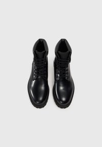 Bottes chelsea en cuir noir à la finition lisse, bout rond, design lacé et col rembourré. Comprend des œillets en métal et une semelle texturée.