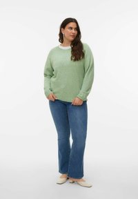 Femme aux longs cheveux bruns portant un pull vert clair, un jean bleu et des chaussures beige à talons bas, debout devant un fond blanc.