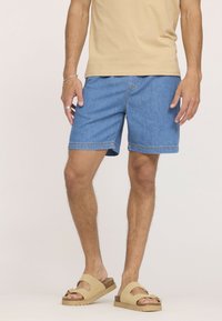 Homme portant un short en denim bleu clair, un t-shirt beige, des sandales beiges, une bague en argent et un bracelet, se tenant devant un fond uni.