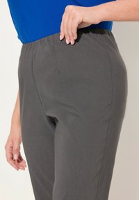Pantalon gris à taille élastique avec une texture lisse. Détail sur la couture latérale visible, associé à un haut bleu à manches courtes, la main reposant sur la taille.