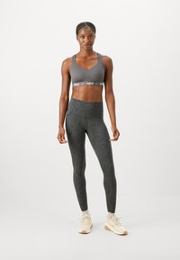 Grauer sportlicher Sport-BH mit breitem Nike-Logo-Band, kombiniert mit hochgeschnittenen schwarzen Leggings, die ein dezentes Wirbelmuster aufweisen, sowie beigen Sneakers.