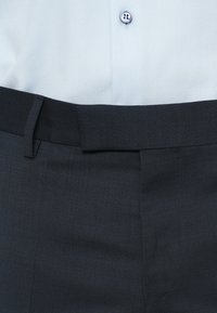 Mörkgrå byxor med en texturerad tyg och en slät frontdesign. Utrustade med en slät svart midjeband och en ljusblå, mönstrad skjorta ovanför.