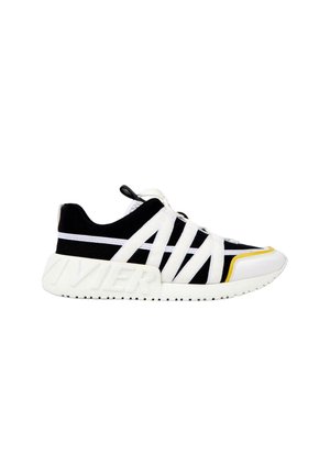 Baskets basses noir et blanc avec des brides croisées blanches, une semelle blanche épaisse avec un logo embossé et un petit accent jaune près des orteils.