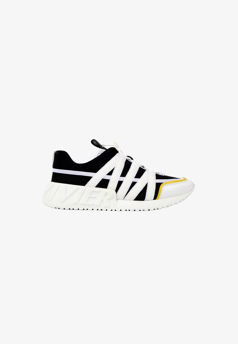 Baskets basses noir et blanc avec des brides croisées blanches, une semelle blanche épaisse avec un logo embossé et un petit accent jaune près des orteils.
