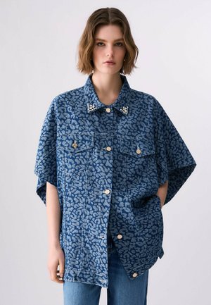 Giovane donna che indossa una giacca di jeans oversize blu con stampa leopardata, colletto decorato e jeans blu, in piedi contro uno sfondo semplice.