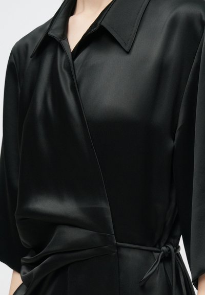 Blusa envolvente de cetim negro com colarinho pontudo, apresentando um detalhe atado na cintura e uma textura suave e brilhante.