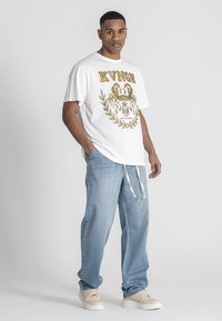 Camiseta gráfica blanca con detalles en dorado, que presenta el texto "KVNGH" y un diseño de escudo; combinada con jeans holgados de color azul claro y zapatillas beige.