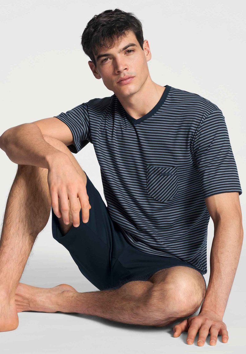 Marineblauwe en grijze gestreepte short-sleeve shirt met een zak en donkere shorts. Model zit, toont huidtextuur en een ontspannen pose.