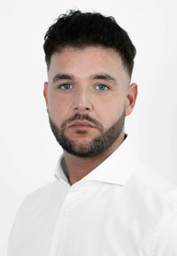 Portret van een persoon met kort, donker haar en een baard, gekleed in een wit overhemd met kraag, tegen een plain witte achtergrond.