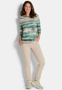 Mehrfarbiger Pullover mit Zickzackmuster in Teal, Weiß und Grau, kombiniert mit hellbeigen Hosen und hellen Sneakers.