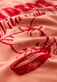 Virsiku värvi puuvillane T-särk punase graafilise kujundusega, kujutades kassi, kes joob kausist, omades sujuvat tekstuuri ja veidi kortsus välimust.