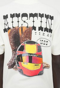 Grafisk t-skjorte med brune cowboy-støvler og en fargerik racinghjelm. Trykt tekst inkluderer "Austin, Texas" og "1025." Hvit stoff.