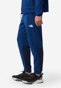 Niebieskie spodnie sportowe z teksturowanymi ciemnoniebieskimi panelami, elastycznym pasem i białym logo The North Face. Połączone z czarnymi butami do biegania.