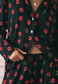 Mustaline komplekt mustades pidžaamades, millel on punased ja kuldsed suudlusprindid. Särgil on krae ja nupud, pükstel on elastne vöökoht.