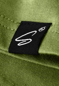 Tessuto verde con una trama texture, caratterizzato da un'etichetta nera cucita sul bordo, che mostra un logo stilizzato bianco.