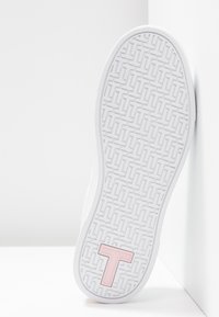 Semelle de sneaker blanche avec motif géométrique texturé et un grand "T" rose près du talon, positionnée sur un fond blanc en coin.