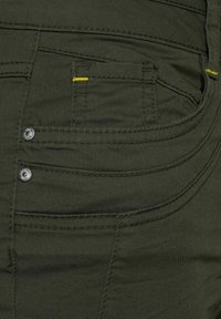 Närbild av en mörkgrön denimbyxas ficka med gul sömnad och två metallnit på kanten.