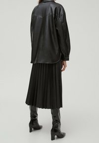 Giacca camicia in pelle nera con colletto a punta, gonna plissettata nera e stivali lucidi al ginocchio con tacco cubano. Texture complessivamente liscia.