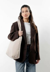 Beige toteväska med en plyschliknande textur, med en subtilt böjd form och ett enda axelband, hålls av en person i en mörk oversized blazer.